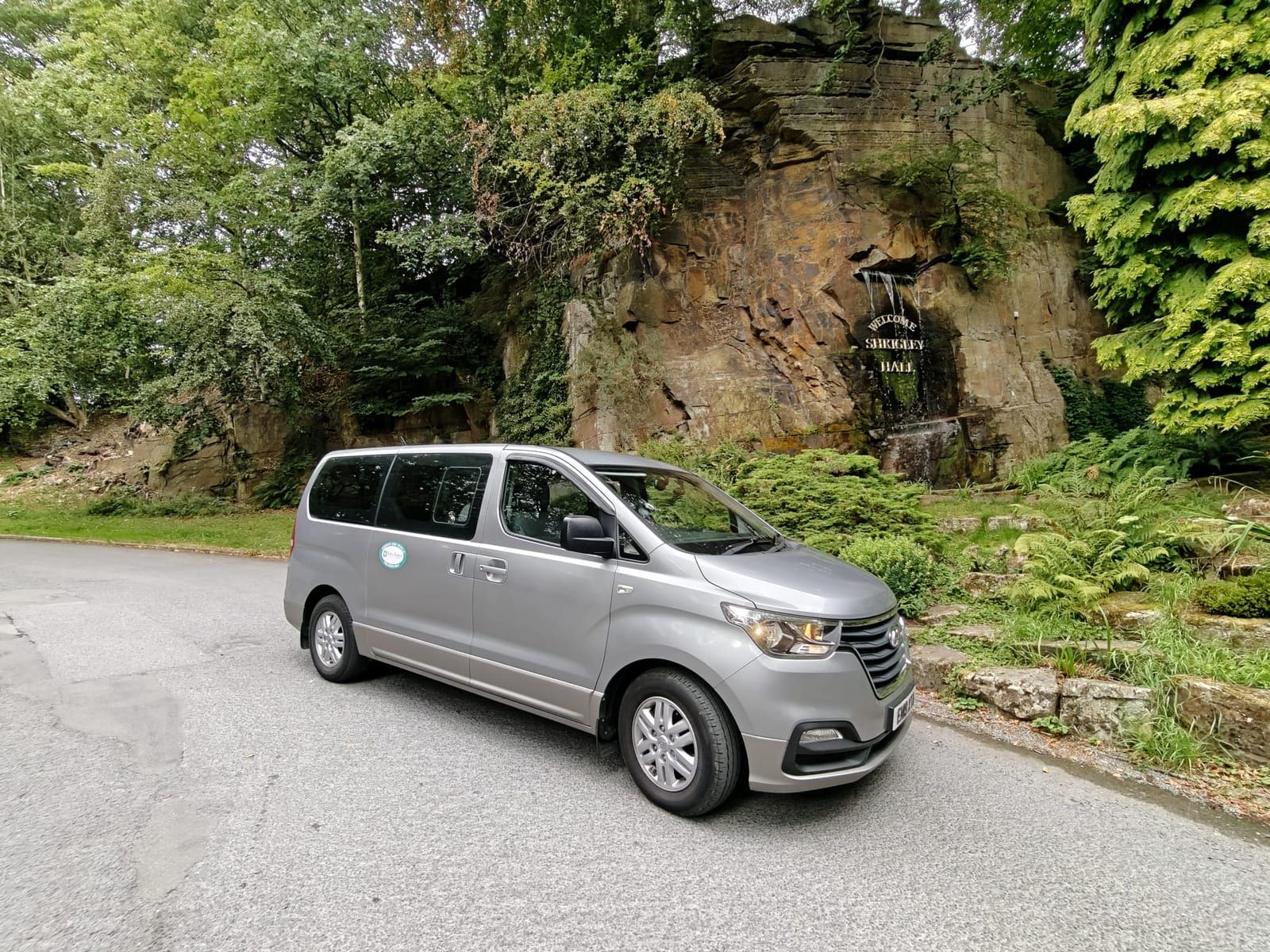 A silver minibus parked on a tree-lined drive beside a rock garden and waterfall.